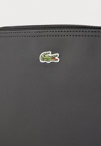Sacca in tessuto nero con texture, caratterizzata da un piccolo logo di un coccodrillo ricamato verde e rosso. Include una cerniera nera lungo il bordo superiore.
