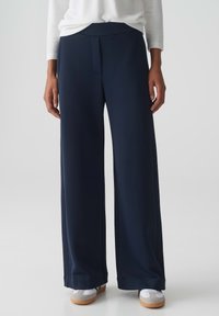 Marineblauwe broek met wijde pijpen en een zachte textuur, gekenmerkt door een voorplooie en een platte tailleband. Gecombineerd met witte en grijze schoenen.