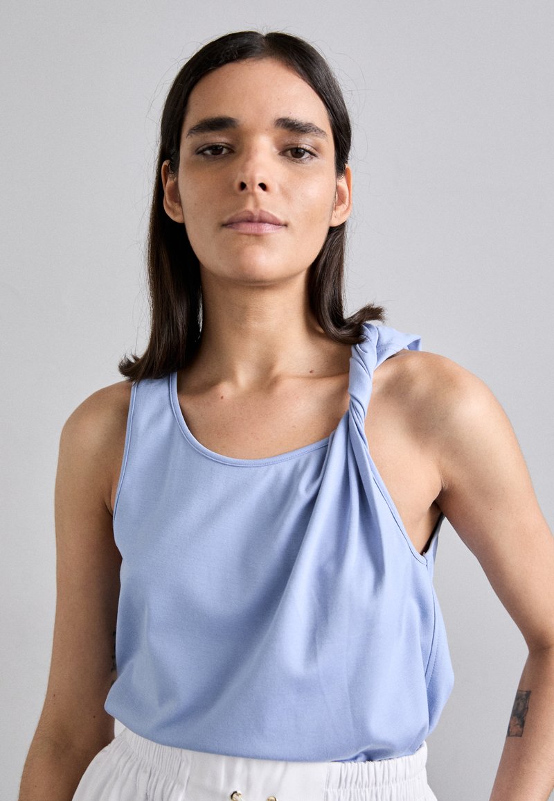 Lichtblauwe mouwloze top met een gedraaide schouderdetail, gemaakt van een gladde stof. Ronde halslijn en een losse pasvorm, gecombineerd met hoge taille broek.