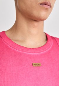 Różowa, prążkowana bawełniana koszulka z okrągłym dekoltem i brązową skórzaną metką z logo "HUGO" wyszytym na klatce piersiowej.