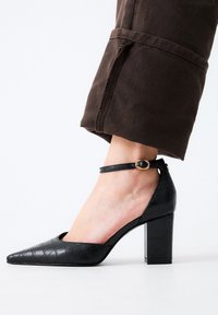 Zapato negro de tacón alto con punta afilada, textura de embossing de cocodrilo, correa en el tobillo con hebilla dorada y tacón grueso.