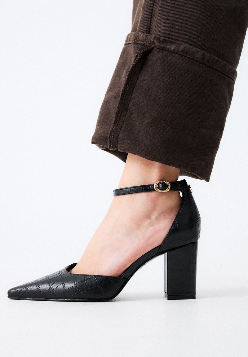 Zapato negro de tacón alto con punta afilada, textura de embossing de cocodrilo, correa en el tobillo con hebilla dorada y tacón grueso.