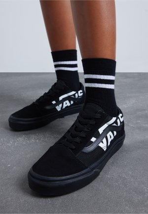 Pies que llevan zapatillas Vans negras con logo blanco y calcetines negros con dos franjas blancas, de pie sobre un suelo de concreto gris.