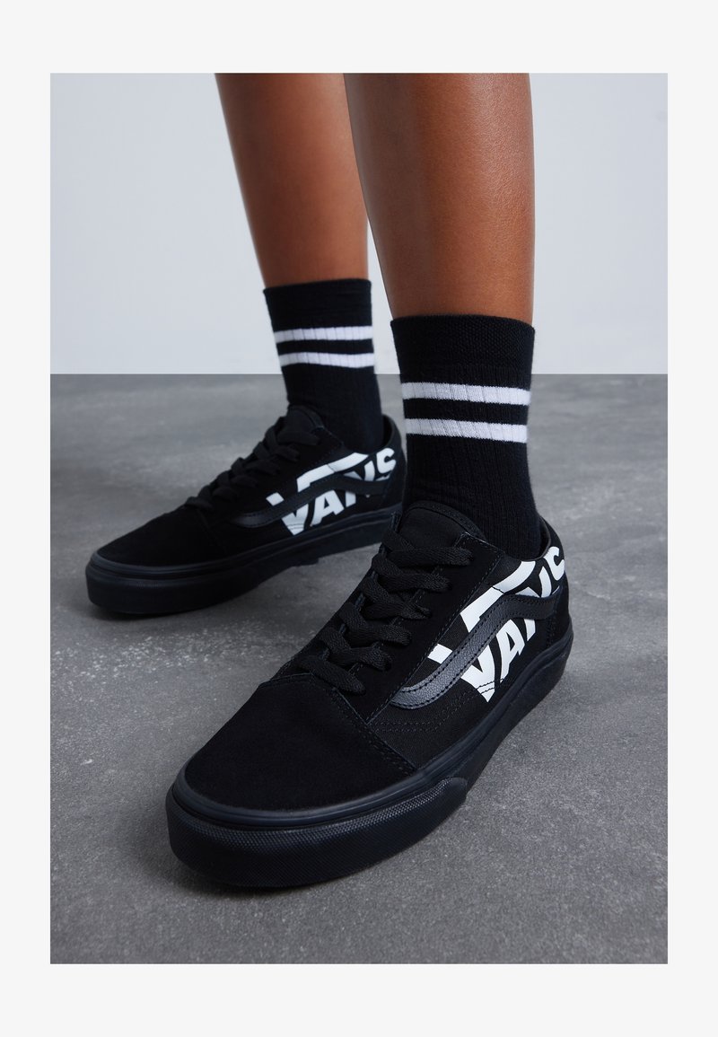 Des pieds portant des baskets Vans noires avec un logo blanc et des chaussettes noires à deux rayures blanches, se tenant sur un sol en béton gris.