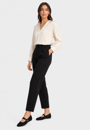 Vrouw draagt een crèmekleurige blouse, zwarte broek met hoge taille en zwarte Mary Jane ballerina's, staand met één hand in de zak tegen een effen achtergrond.