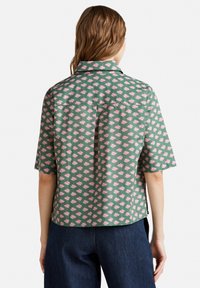 Donna che indossa una maglietta verde a maniche corte con motivo floreale rosa, abbinata a jeans blu scuro, vista da dietro su uno sfondo bianco.