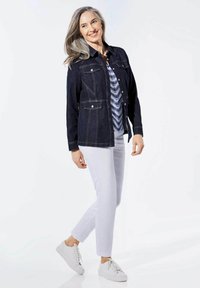 Dunkelblaue Jeansjacke mit zwei Brusttaschen und weißen Nähten, kombiniert mit einem blauen gemusterten Oberteil und weißen Hosen. Getragen mit weißen Sneakers.