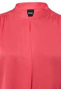 Koraalroze blouse met opstaande kraag en keyhole halslijn, gladde stof, geplooide schouders en zwart "BOSS" label binnenin de kraag.