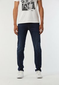 Jean en denim bleu foncé avec une coupe slim, doté d'un design classique à cinq poches. Associé à un t-shirt graphique blanc et des baskets blanches.