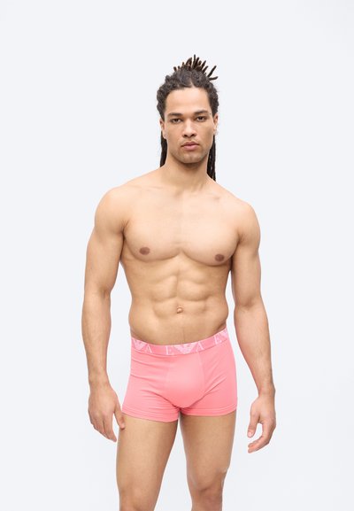 Μυώδης άνδρας με ντραντλοκς στέκεται μπροστά σε λευκό φόντο, φορώντας ροζ μποξεράκια με λογότυπο στη μέση, κοιτάζοντας μπροστά.