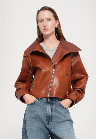 Giacca cropped in pelle marrone con ampi revers, chiusura lampo frontale e tasche laterali, abbinata a jeans chiari a vita alta.