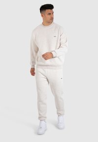 Sudadera gris claro y joggers a juego hechos de una tela suave y texturizada. Presenta un pequeño logo en el pecho y en la parte superior de la pierna, complementado por zapatillas blancas.