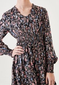 Czarna sukienka w kwiaty z różowymi, niebieskimi i kremowymi wzorami. Posiada talię z marszczeniem, falbaniasty dekolt oraz długie rękawy z marszczonymi mankietami.