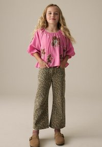 Haut rose avec broderie florale, coupe ample, manches larges ; associé à un pantalon à jambes larges en imprimé léopard ; sabots bruns avec détail de boucle.