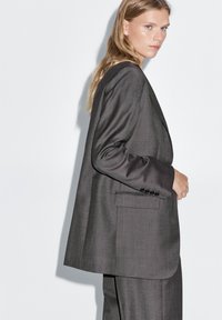 Gray tailored suit jacket with a structured silhouette, notched lapels, and a smooth fabric texture. Features side pocket and buttoned cuff.
