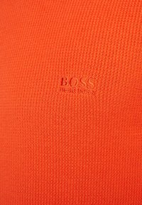 Orangener strukturierter Stoff mit subtiler Strickmuster. Prägiertes Logo "BOSS" und "HUGO BOSS" in passender Farbe unten rechts.