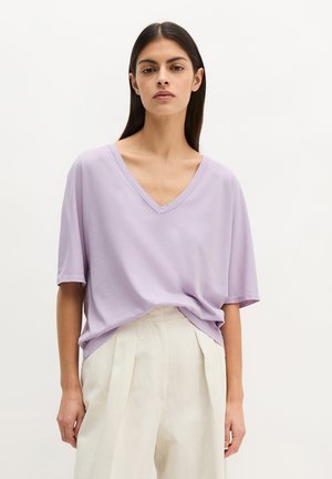 Vrouw draagt een losse lila V-hals top met korte mouwen, ingestopt in een hooggetailleerde, crèmekleurige geplooide broek, staand tegen een effen achtergrond.