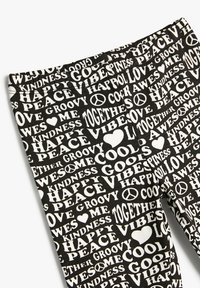 Leggings negros con un patrón de texto en blanco que incluye palabras como "PAZ", "AMOR" y "FELIZ", junto con signos de paz y corazones.