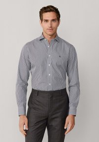 Pitkähihainen nappikaulusm desprouse, jossa on tummansinisiä ja valkoisia pystyraitoja, puuvillakangasta sekä pieni logo rinnassa; yhdistetty tummanharmaiden housujen kanssa.