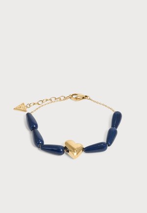 Bracelet chaîne en or avec un pendentif cœur en or au centre et six perles en forme de goutte bleu foncé, avec une petite étiquette triangulaire en or.