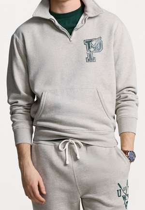 Uomo che indossa una felpa con zip parziale grigio chiaro e pantaloni della tuta coordinati con loghi verdi, orologio al polso sinistro e maglietta verde sotto.