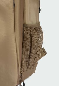 Beżowy plecak z teksturowaną tkaniną, siatkową boczną kieszenią oraz detalami na zamek, o gładkim i wytrzymałym wykończeniu.