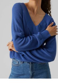 Maglione blu lavorato a maglia con scollo a V, maniche lunghe e polsini a coste. Indossato con jeans blu a vita alta, che mostrano un motivo strutturato.