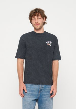 Homme aux cheveux bruns bouclés portant un t-shirt foncé délavé avec un petit texte "Aranciata" et un motif fruité sur la poitrine, ainsi qu'un jean bleu clair.