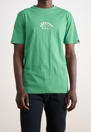 Vihreä puuvillapaita, jossa on vaalean kermanvärinen etulogo, lyhyet hihat ja pyöreä kaula-aukko, yhdistettynä mustiin housuihin.