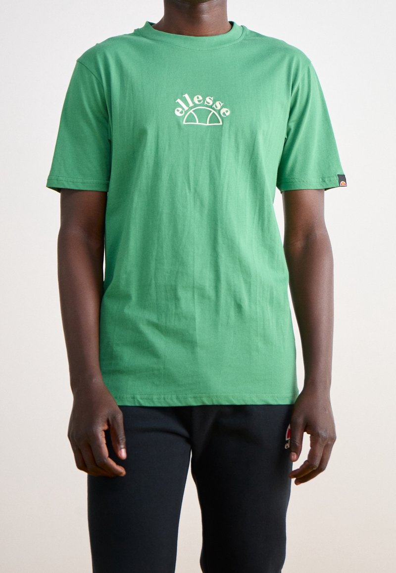 Camiseta de algodón verde con un diseño de logo en crema en la parte delantera, mangas cortas y cuello redondo, combinada con pantalones negros.