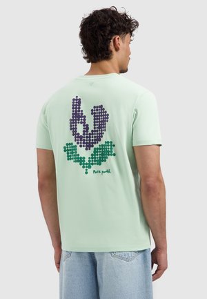 Man met krullend haar draagt een lichtgroen T-shirt met een paars-groen gestippeld bloemontwerp en de tekst "Pure path" op de rug.