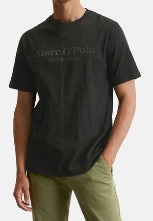 Mann trägt ein schwarzes Marc O'Polo T-Shirt mit kurzen Ärmeln und olivgrüner Hose, mit einer Hand in der Tasche vor weißem Hintergrund.