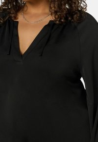 Svart blus med V-ringning, dragsko-detaljer och långa ärmar. Slät, lätt tyg med avslappnad passform.