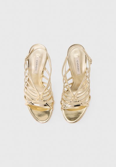 Χρυσά σανδάλια με ψηλό τακούνι και λουράκια, με κομψό μεταλλικό φινίρισμα, ανοιχτό δάχτυλο και λουράκια που διασταυρώνονται. Διαθέτουν εσωτερική σόλα με λογότυπο.