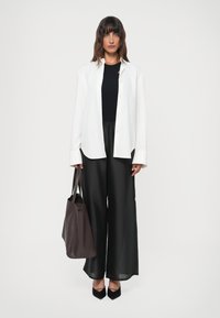 Woman standing wearing a white buttoned shirt over a black top and wide black pants, holding a dark brown large tote bag, black heels.