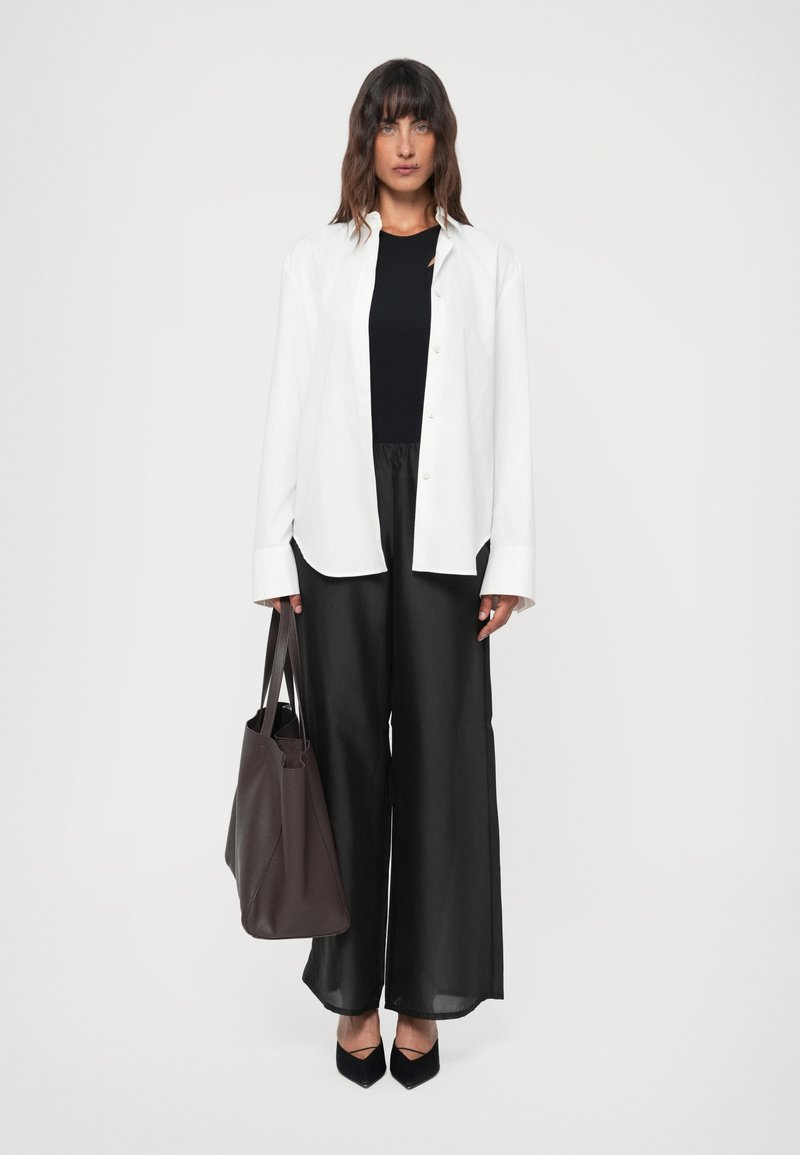 Woman standing wearing a white buttoned shirt over a black top and wide black pants, holding a dark brown large tote bag, black heels.