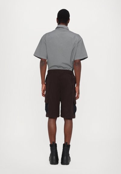 Chemise grise à manches courtes avec un col, associée à un short cargo marron foncé. Des bottes noires à semelle haute complètent la tenue.