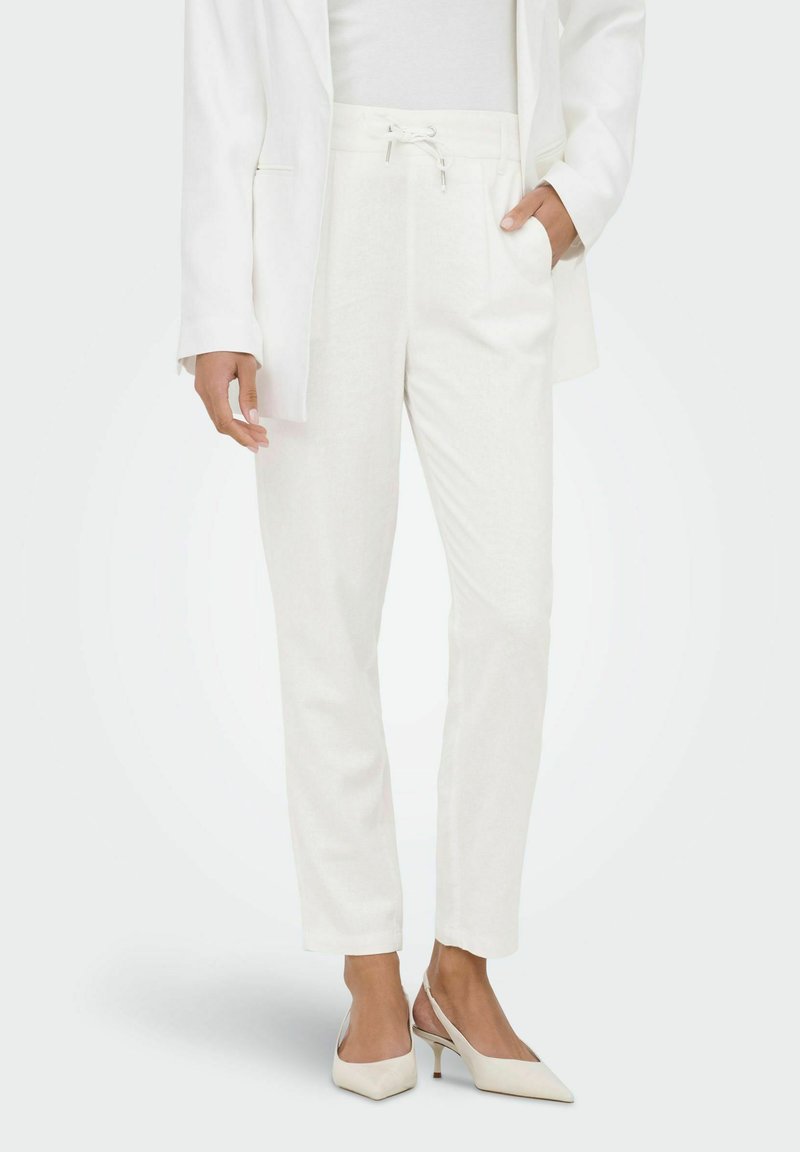 Femme portant un pantalon blanc sur mesure à cordon, un blazer blanc et des talons blancs à bride arrière, debout avec une main dans la poche.