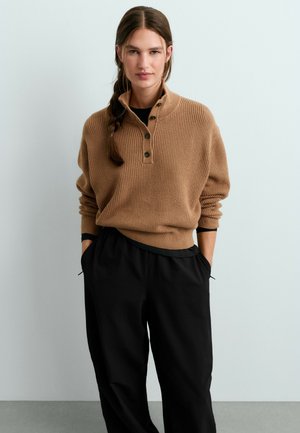 Femme aux cheveux tressés portant un pull en maille marron boutonné et un pantalon noir à jambes larges, debout les mains dans les poches sur un fond clair.