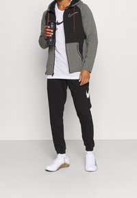 Haut à capuche zippé gris et noir, t-shirt blanc, joggeurs noirs et baskets blanches avec semelles en caoutchouc. Tient une bouteille d'eau noire.