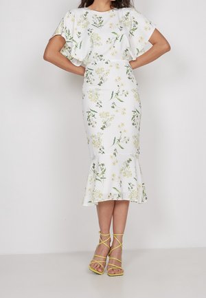 Woman wearing a white floral midi dress with ruffled hem and flutter sleeves, paired with yellow strappy high-heeled sandals.