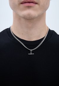 Strieborný náhrdelník z reťaze s hrubým prepojením a vodorovným prútikom, nosený cez jednoduché čierne tričko.