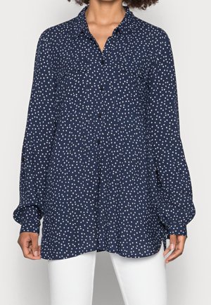 Kobieta ubrana w granatową bluzkę na guziki z długim rękawem w białe kropki oraz białe spodnie, stojąca na jednolitym tle.