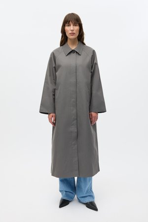 Long manteau gris avec un grand col, fermeture boutonnée à l'avant et manches longues ; associé à un jean bleu à jambes larges et des chaussures noires à bout pointu.