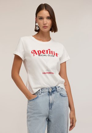 Kvinde med kort brunt hår iført store sølvøreringe, en hvid t-shirt med rød tekst "Aperitif Social Club Happiness," og lyseblå jeans.