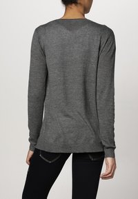 Osoba w długim szarym swetrze i czarnych spodniach widoczna od tyłu na jednolitym tle.