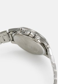 Montre-bracelet en métal argenté avec un bracelet à maillons, montrant le côté avec deux boutons-poussoirs et une couronne sur fond blanc.