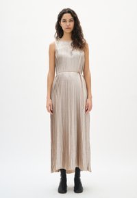 Metaliczna plisowana sukienka w jasnobeżowym kolorze z okrągłym dekoltem, taliowaną talią i długością do kostek. Gładka faktura, zestawiona z czarnymi botkami.