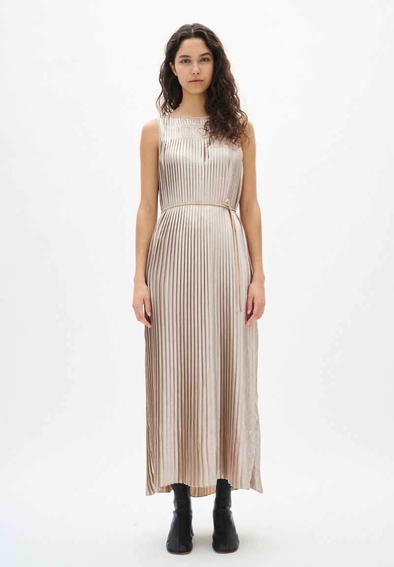 Metaliczna plisowana sukienka w jasnobeżowym kolorze z okrągłym dekoltem, taliowaną talią i długością do kostek. Gładka faktura, zestawiona z czarnymi botkami.