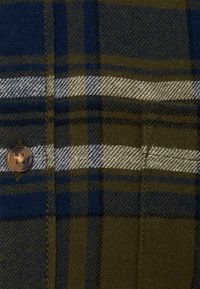 Tissu en mélange de laine avec un motif à carreaux vert, marine et blanc ; présente un bouton marron sur le côté gauche. La texture semble douce et texturée.